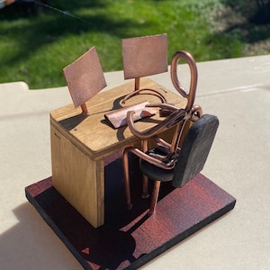 May include: A handcrafted sculpture of a person at a desk, made from copper wire and wood. The desk has two copper monitor screens and a small wooden chair. The base is a dark stained wood.