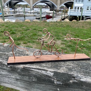May include: A copper wire sculpture of three figures on a wooden base. One figure is riding a bicycle, another is running, and the third is swimming.