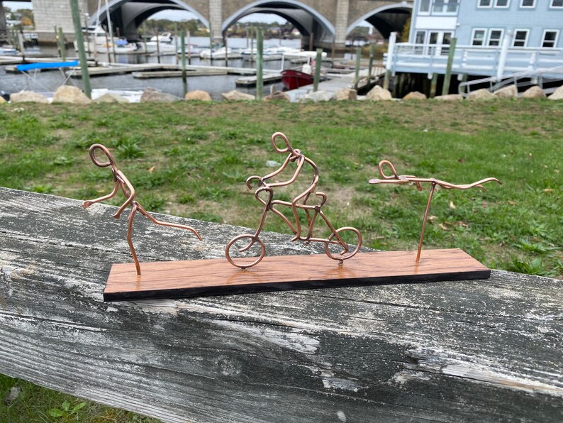 May include: A copper wire sculpture of three figures on a wooden base. One figure is riding a bicycle, another is running, and the third is swimming.