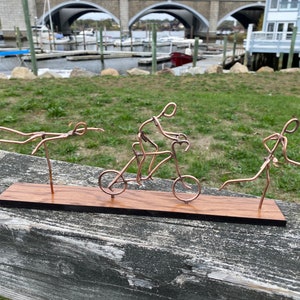 May include: A wooden base with three wire sculptures depicting a swimmer, a cyclist, and a runner. The sculptures are made of copper wire and are arranged in a line on the base.