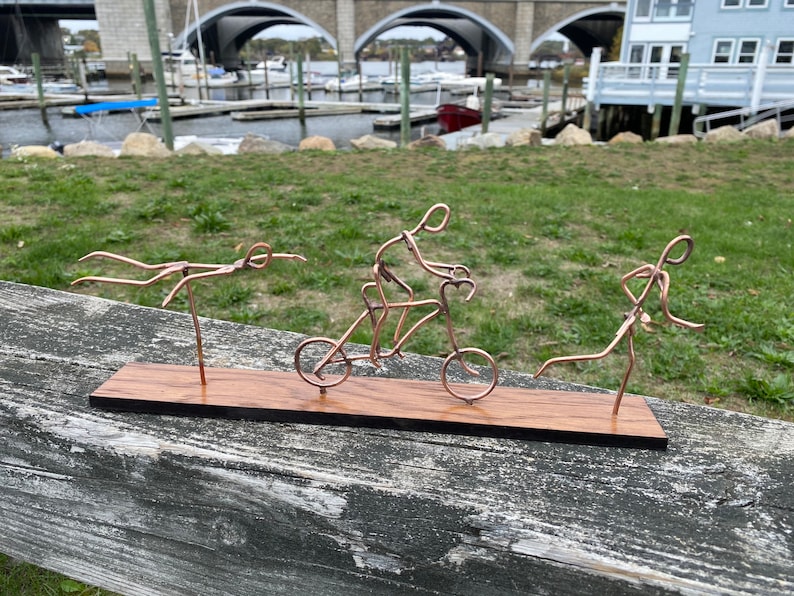 May include: A wooden base with three wire sculptures depicting a swimmer, a cyclist, and a runner. The sculptures are made of copper wire and are arranged in a line on the base.