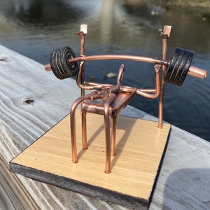 May include: A copper-colored metal sculpture of a weight bench with a barbell. The bench is on a wooden base with a dark brown border.