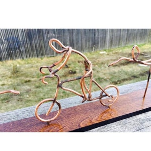 May include: A copper wire sculpture of a person riding a bicycle. The sculpture is mounted on a wooden base.