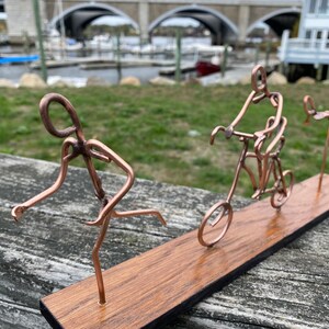 May include: A copper wire sculpture of three figures on a wooden base. The figures are a runner, a person riding a bicycle, and a person on a unicycle.