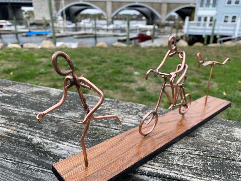 May include: A copper wire sculpture of three figures on a wooden base. The figures are a runner, a person riding a bicycle, and a person on a unicycle.