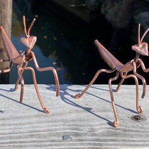 May include: Two copper praying mantis sculptures, each with a detailed body and six legs, are posed on a wooden surface.
