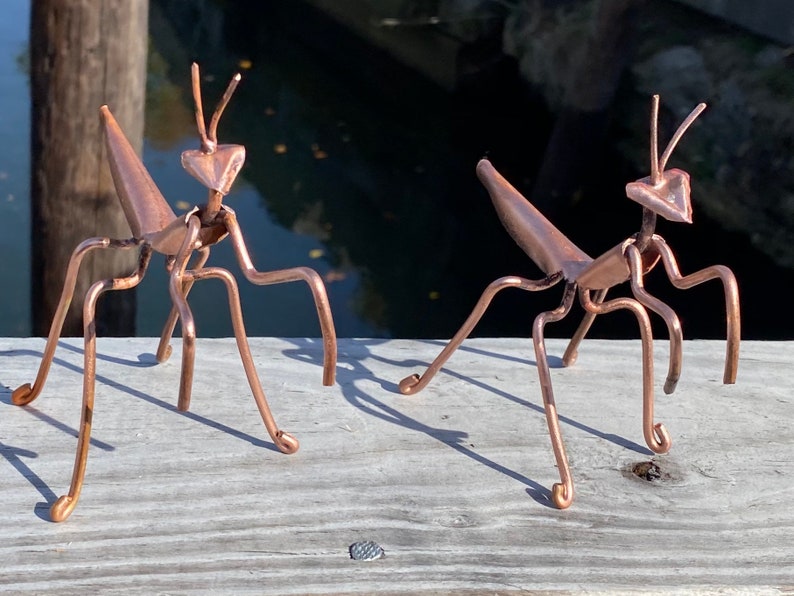 May include: Two copper praying mantis sculptures, each with a detailed body and six legs, are posed on a wooden surface.