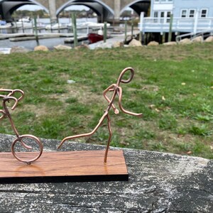 May include: A copper wire sculpture of two figures, one riding a bicycle and the other running. The sculpture is mounted on a wooden base.