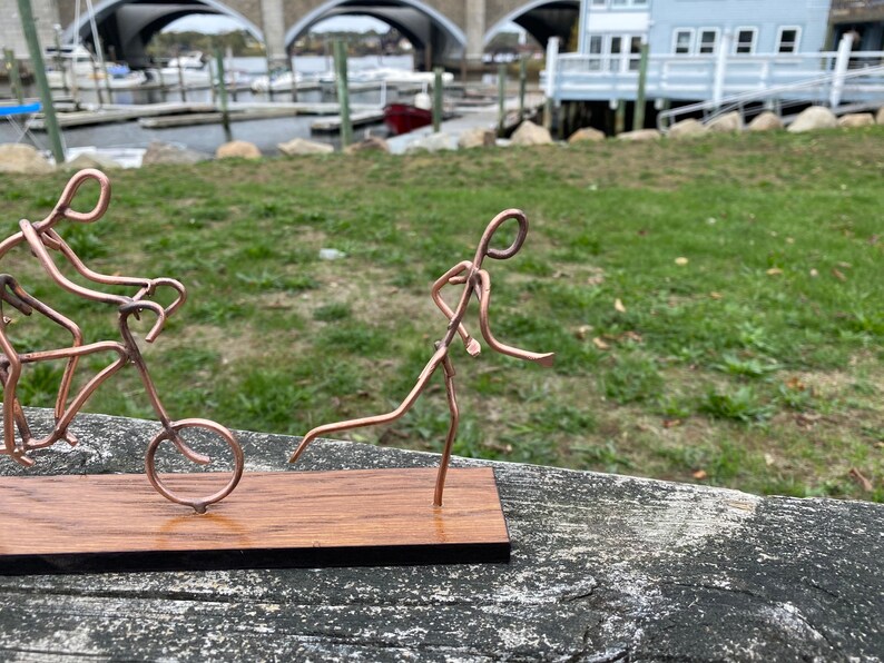 May include: A copper wire sculpture of two figures, one riding a bicycle and the other running. The sculpture is mounted on a wooden base.