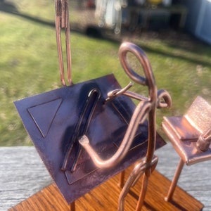 May include: A copper-colored metal sculpture of a person sitting at a desk with a lamp and a chair. The person is drawing on a piece of paper with a ruler. The sculpture is on a wooden base.