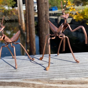 May include: Two copper-colored metal praying mantis sculptures on a wooden surface. The sculptures are detailed and realistic.