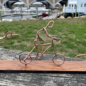 May include: A copper wire sculpture of three figures on a wooden base. One figure is riding a bicycle, another is doing a handstand, and the third is standing with one leg raised.