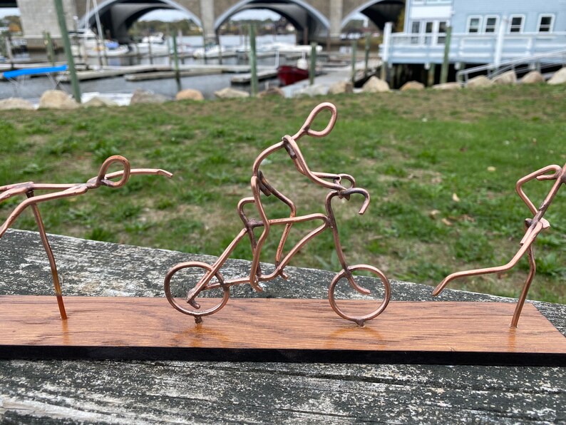 May include: A copper wire sculpture of three figures on a wooden base. One figure is riding a bicycle, another is doing a handstand, and the third is standing with one leg raised.