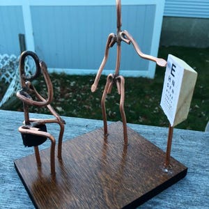 May include: A copper wire sculpture of two figures, one holding an eye chart, on a wooden base. The eye chart reads "E H N D F Z P N Z".