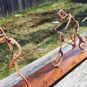 May include: A copper wire sculpture of three figures on a wooden base. The figures are a person running, a person riding a bicycle, and a person diving. The sculpture is a unique and creative piece of art.
