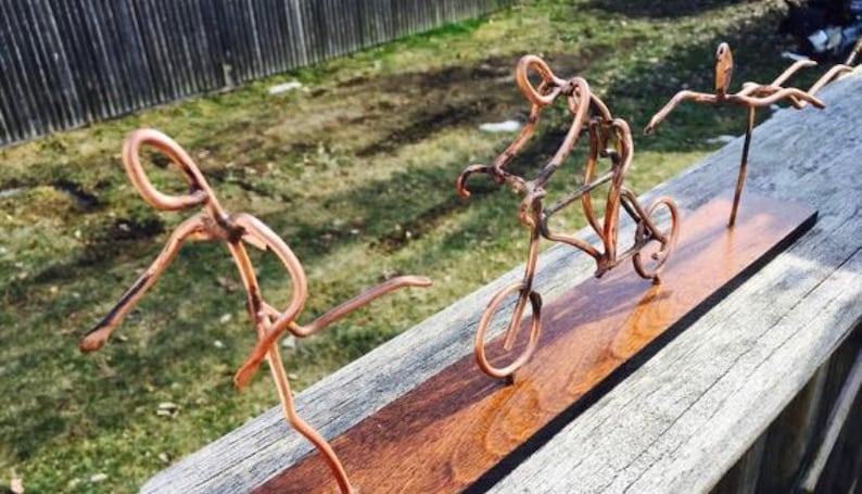 May include: A copper wire sculpture of three figures on a wooden base. The figures are a person running, a person riding a bicycle, and a person diving. The sculpture is a unique and creative piece of art.