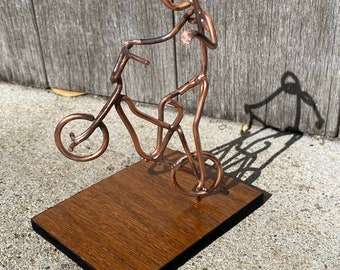 Op de afbeelding: Een sculptuur van koperdraad van een persoon die op een fiets rijdt op een houten basis. Het beeld is gemaakt van dun draad en is zeer gedetailleerd.