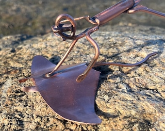 Op de afbeelding: Een koperkleurig metalen beeldje van een duiker die op een mantarog rijdt. Het beeldje staat op een ruw, bruin rotsoppervlak.