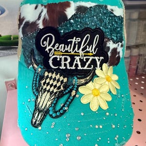 May include: A turquoise baseball cap featuring a cow print patch, a black embroidered patch with the text Beautiful CRAZY, a bull skull graphic with turquoise accents, two yellow daisy appliques, and scattered clear rhinestones on the brim.