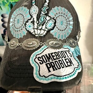 May include: Gray baseball cap with decorative patches and a silver chain. The patches include a peace sign, a lightning bolt, and the phrase "SOMEBODY'S PROBLEM" in white lettering with a turquoise border. The cap has a distressed look.