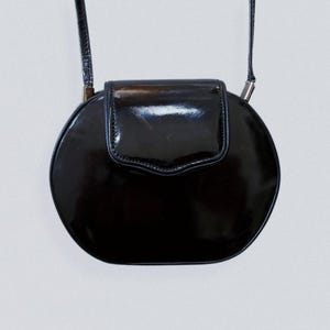 May include: A black, round, patent leather crossbody bag with a flap closure. The bag has a thin strap and silver-toned hardware. The glossy finish reflects light, highlighting its smooth surface. The bag's design is simple and elegant.