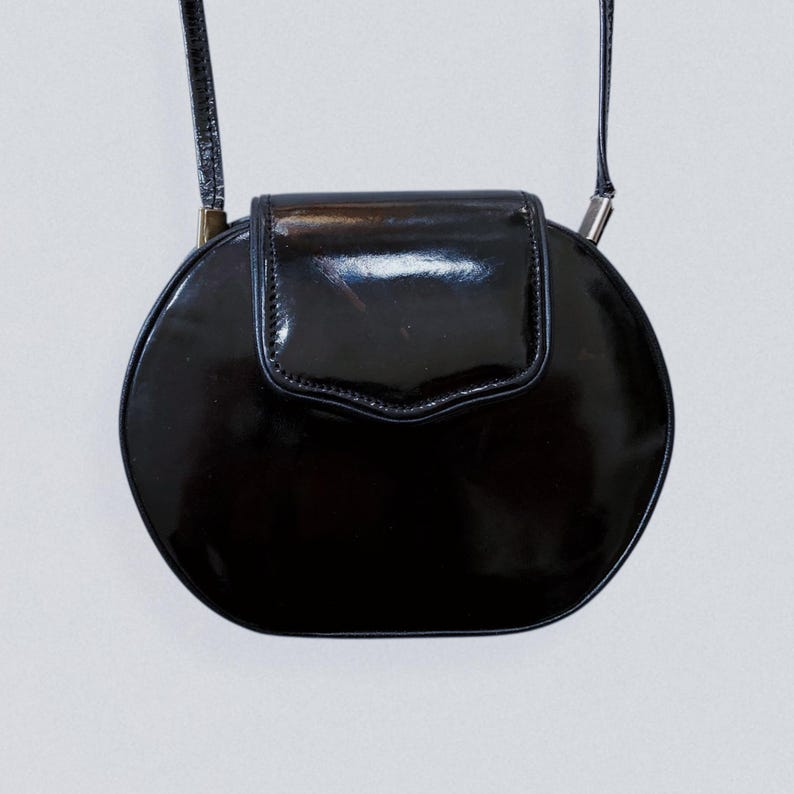 May include: A black, round, patent leather crossbody bag with a flap closure. The bag has a thin strap and silver-toned hardware. The glossy finish reflects light, highlighting its smooth surface. The bag's design is simple and elegant.