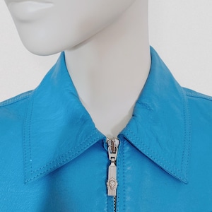 May include: A close-up of a turquoise leather jacket with a silver zipper. The jacket has a classic collar and is displayed on a mannequin. The zipper pull has a decorative emblem.