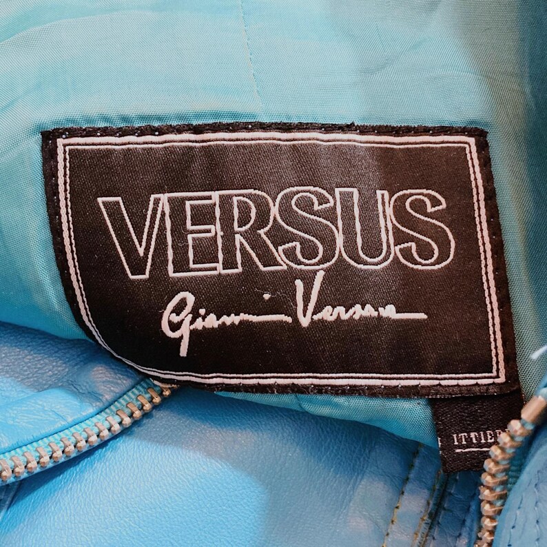 May include: Close-up of a turquoise leather jacket with a silver zipper. The black label reads "VERSUS" in white capital letters, with "Gianni Versace" in cursive below. The jacket's stitching and texture are visible.