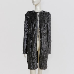 Peut inclure: Un long manteau noir avec un design texturé et superposé. Le manteau a un col rond et une fermeture à nouer. Le vêtement est présenté sur un mannequin sur fond blanc. La texture du manteau lui donne un aspect unique et accrocheur.