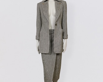 May include: A gray tweed skirt suit with a long blazer and a knee-length skirt. The blazer has a single button closure and flap pockets. The skirt has a wrap design.