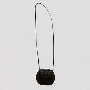 May include: A small, black, round crossbody bag with a glossy finish. The bag features a flap closure and a thin, black strap. The design is simple and elegant, suitable for various occasions.