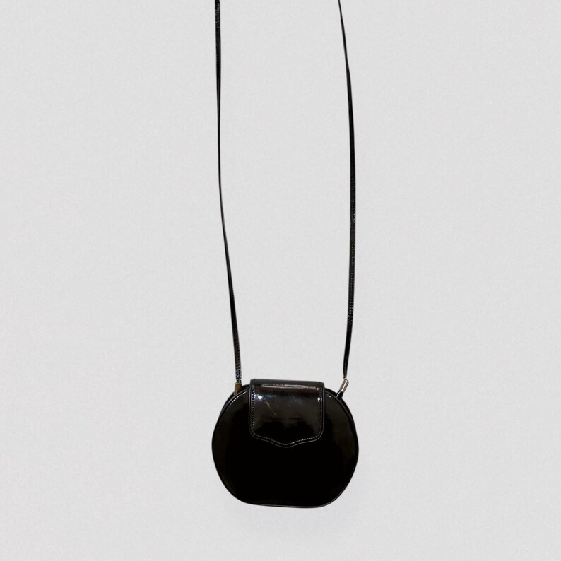 May include: A black, round crossbody bag with a glossy finish. The bag features a flap closure and a long, thin strap. The design is simple and elegant, suitable for various occasions.