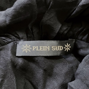 May include: Close-up of a black garment with a label that reads "PLEIN SUD" in gray lettering. The label has a starburst design on each side. The fabric appears to be soft and flowing.