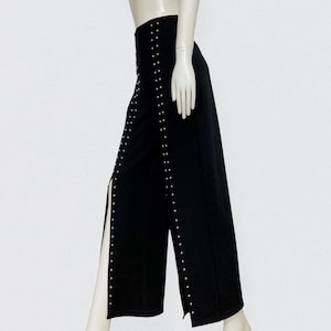 Peut inclure: Pantalon noir à jambes larges avec une taille haute et des fentes latérales. Le pantalon est orné de clous argentés le long des coutures et des fentes. Le pantalon est présenté sur un mannequin.