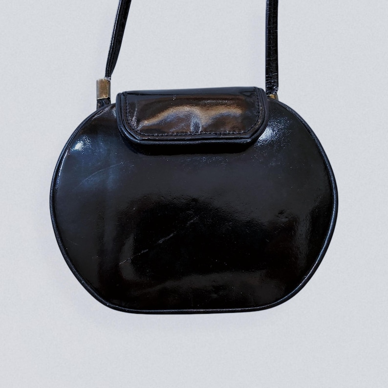 May include: A black, circular crossbody bag with a glossy finish. The bag features a flap closure and a thin strap. The design is simple and elegant, suitable for various occasions. The bag's shape is reminiscent of a half-moon.