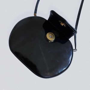 May include: A black, oval-shaped crossbody bag with a glossy finish. The bag features a gold-colored clasp and a thin, black strap. The top flap is open, revealing the interior.