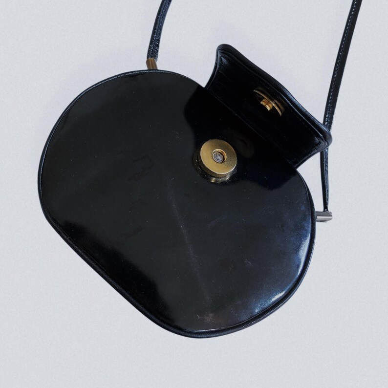 May include: A black, oval-shaped crossbody bag with a glossy finish. The bag features a gold-colored clasp and a thin, black strap. The top flap is open, revealing the interior.