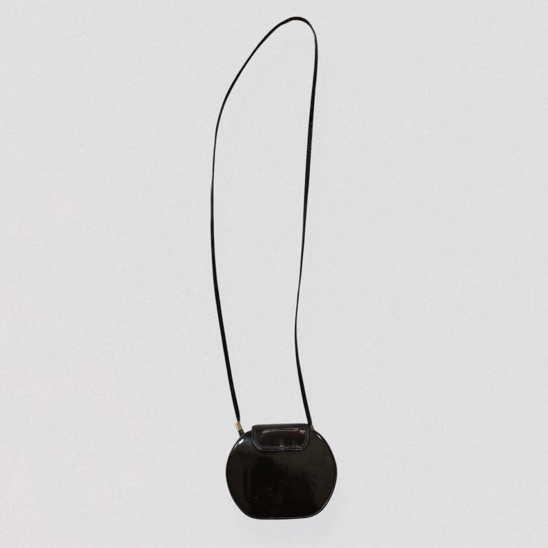 May include: A black, oval-shaped crossbody bag with a long, thin strap. The bag has a glossy finish and a small, rectangular clasp. The strap is attached to the bag with small gold-colored hardware.