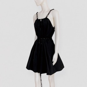 Könnte beinhalten: Ein schwarzes ärmelloses Minikleid mit gerafftem Ausschnitt und einer durch ein schmales Band betonten Taille. Das Kleid ist mit schwarzen Kettenglied-Trägern und einem passenden Kettendetail versehen, das mittig an der Taille herabhängt.