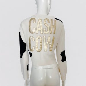 Peut inclure: Pull en laine blanc avec un imprimé vache noir et blanc et un lettrage doré qui dit "CASH COW" au dos.