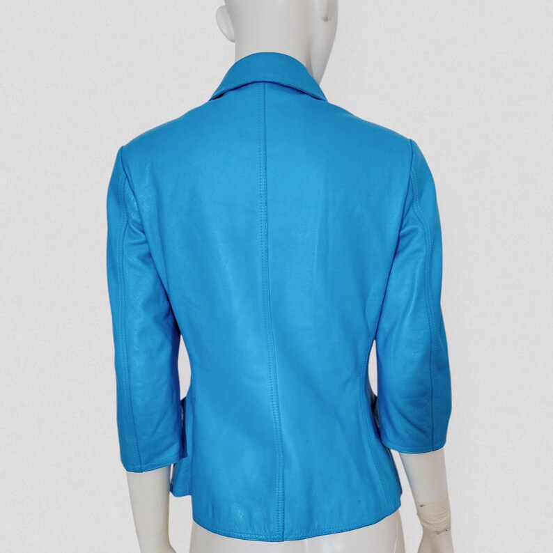 May include: A bright turquoise leather jacket with three-quarter sleeves and a notched collar. The jacket is displayed on a mannequin, showcasing its tailored fit and design. The back view highlights the seams and structure of the garment.