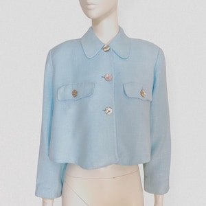 Peut inclure: Veste courte bleu clair avec col, deux poches avant et trois boutons. La veste est présentée sur un mannequin. Les boutons sont de couleur or clair.