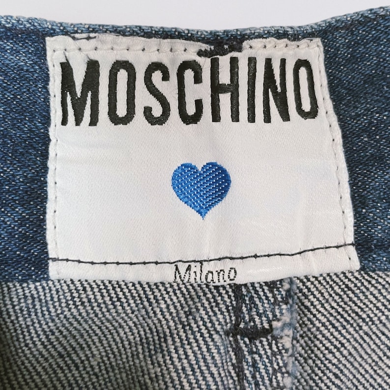 Pode incluir: Uma etiqueta branca com costuras pretas e o texto "MOSCHINO" em letras pretas. Um cora&ccedil;&atilde;o azul com costuras brancas est&aacute; centralizado abaixo do texto. O texto "Milano" est&aacute; centralizado abaixo do cora&ccedil;&atilde;o. A etiqueta est&aacute; presa a um tecido de sarja azul.