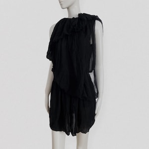May include: Black one-shoulder dress with layered fabric and a ruffled neckline. The dress has a loose, flowing silhouette and appears to be made of a lightweight material. The dress is displayed on a mannequin.