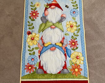 May include: A stack of three gnomes with white beards and red hats, standing on a green patch of grass. The gnomes are surrounded by yellow, red, and blue flowers and green leaves. There are also butterflies and bees flying around the gnomes.