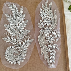 May include: Two silver beaded appliques on a sheer fabric background. One applique features a floral design with multiple flowers and leaves. The other applique features a leafy design with multiple leaves.