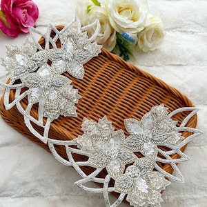 Peut inclure: Deux appliques florales argentées avec des détails complexes de perles et de paillettes. Les appliques présentent un design superposé avec de multiples formes de fleurs et des feuilles ajourées. Elles sont posées sur un panier en osier brun.