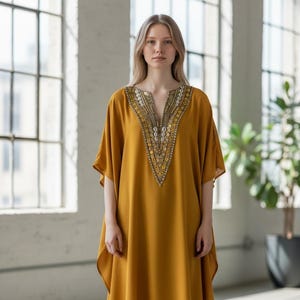 May include: Mustard yellow tunic dress with a V-neckline, embellished with gold and silver details. The dress has short sleeves and a flowing silhouette. The person is wearing blue jeans and white sneakers.