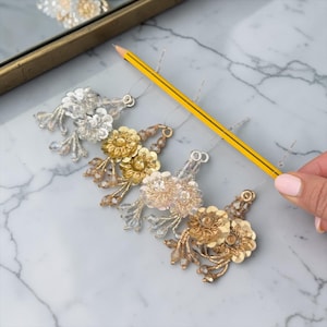 Puede incluir: Primer plano de cuatro accesorios florales para el cabello en tonos plateados, dorados y rosados. Cada pieza presenta detalles de cuentas y lentejuelas. Un lápiz amarillo se coloca junto a los accesorios para la escala. Fondo de mármol.