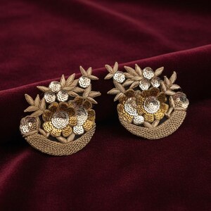 May include: A pair of embroidered floral patches featuring gold and silver metallic thread and sequins. Each patch displays a crescent-shaped base with detailed leaf patterns and circular flower motifs, crafted with intricate needlework and shimmering accents.